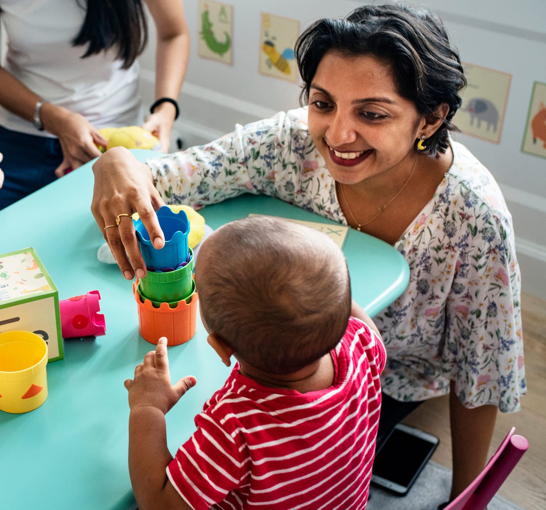 A Creative Playrooms teacher smiling with a toddler stacking colorful cups at a classroom table