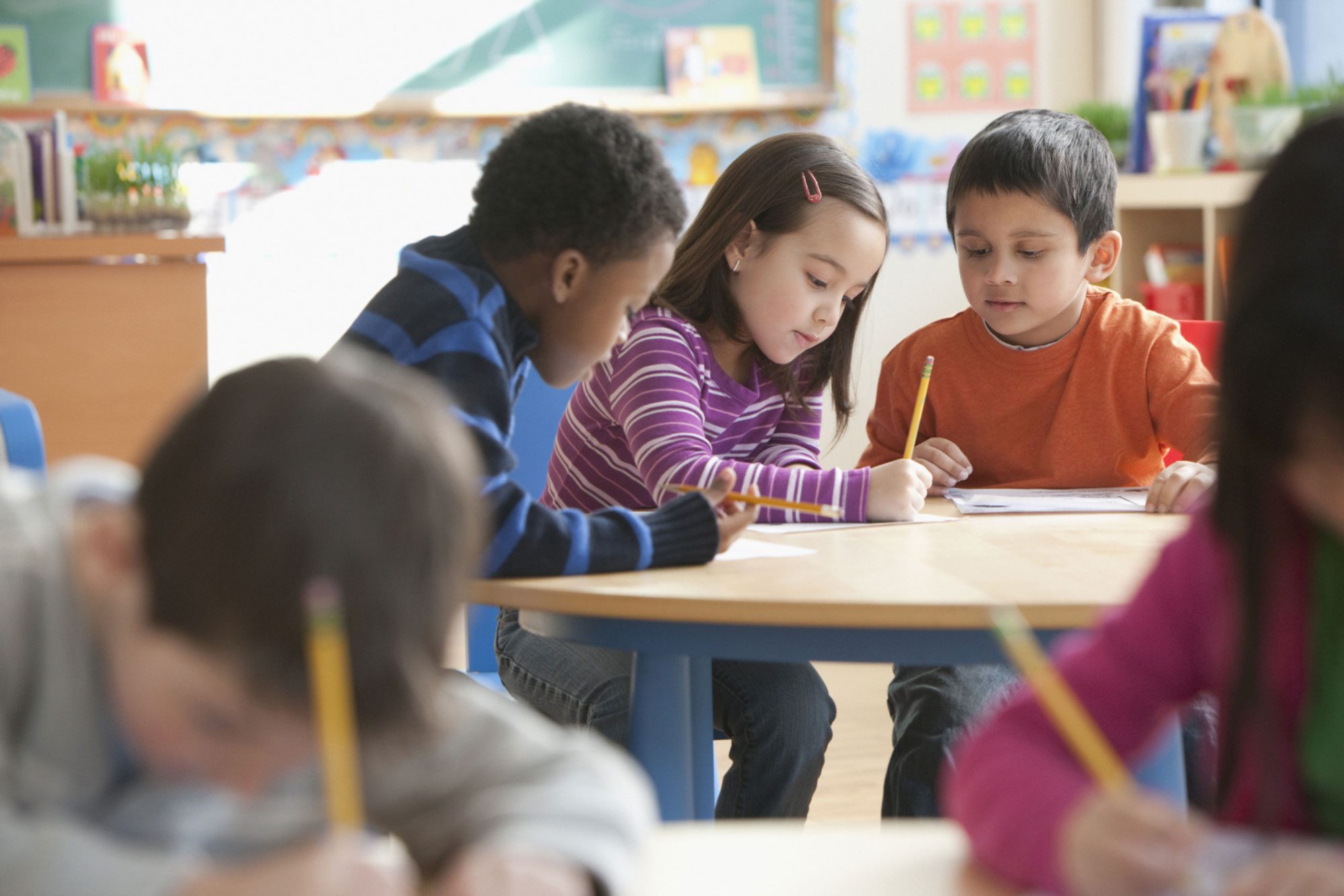 Three children at a classroom table with paper and pencil.